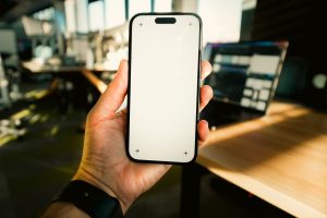 Close-up of hand holding smartphone with blank screen in a sunny office setting.
